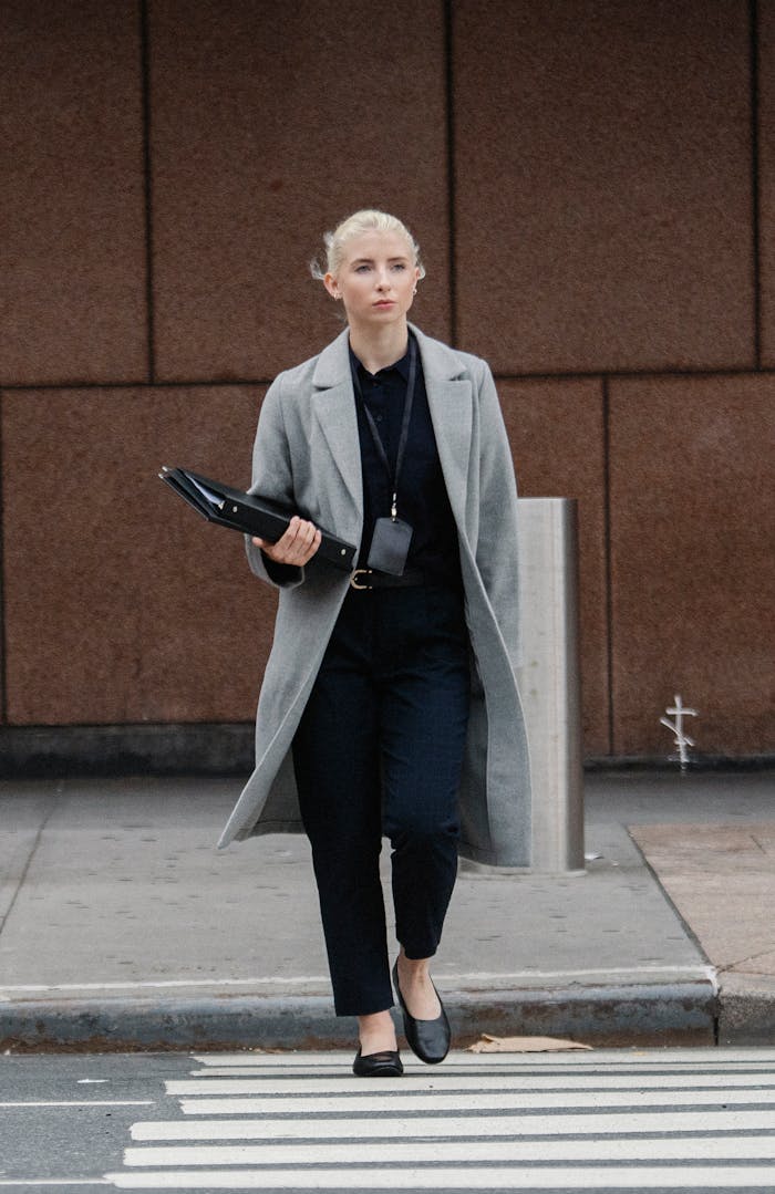 Full body confident young businesswoman in elegant formal wear carrying document folders and crossing road in downtown while looking away thoughtfully